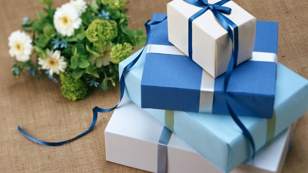 چند جعبه کادوی سفید و آبی در سایزهای مختلف روی هم کنار یک دسته گل سفید