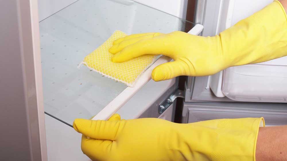 تمیز کردن قفسه‌های شیشه‌ای درون یخچال با اسفنج و دستکش زرد