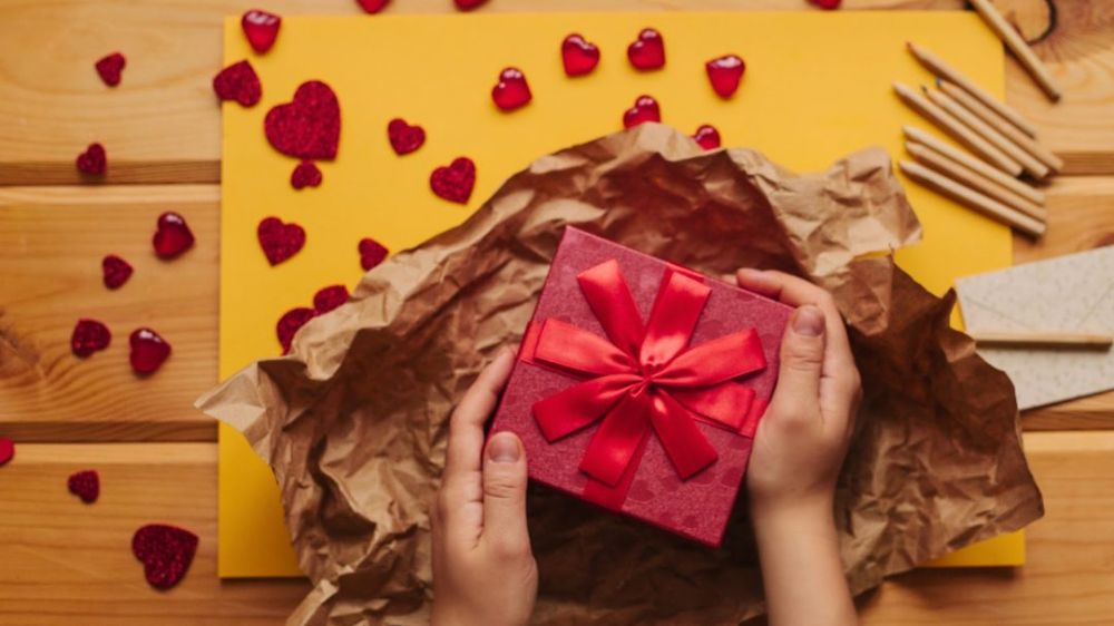 یک دست که یک جعبه هدیه قرمز رنگ را گرفته به همراه چند قلب کوچک روی میز