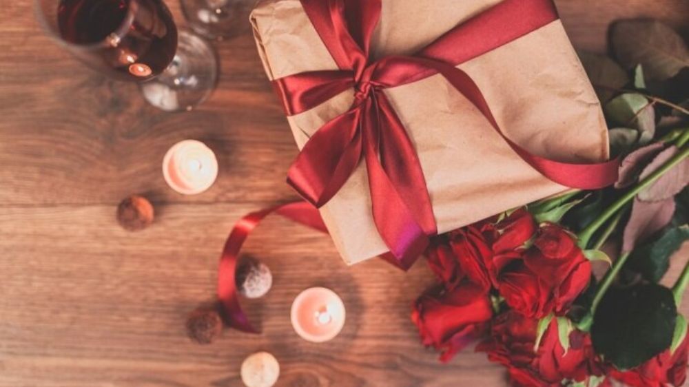 جعبه هدیه بسته بندی شده با روبان قرمز به همراه چند گل و شمع روی یک میز قهوه‌ای