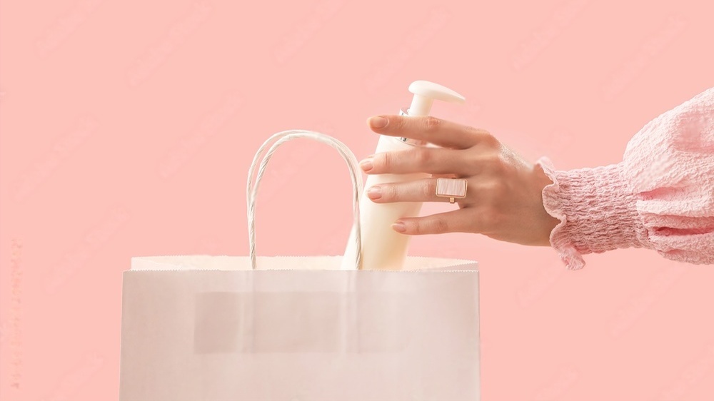 تصویر دست یک خانم در حال بیرون آوردن یک پرایمر سفید رنگ از یک بسته خرید سفید با پس زمینه صورتی