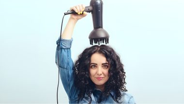خانمی با موهای فر بلند که سشوار و دیفیوزر آن را بالای سرش نگه داشته است.
