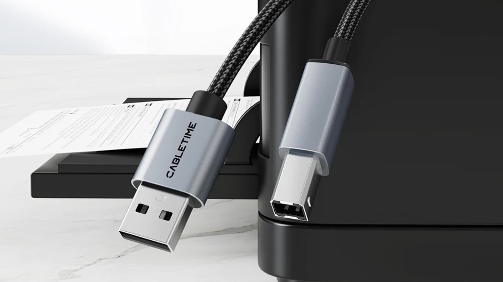 یک کابل USB-A به USB-B بافته شده مشکی با سری‌های متالیک و یک پرینتر مشکی در پشت سر آن روی پس‌زمینه سفید ماربلی