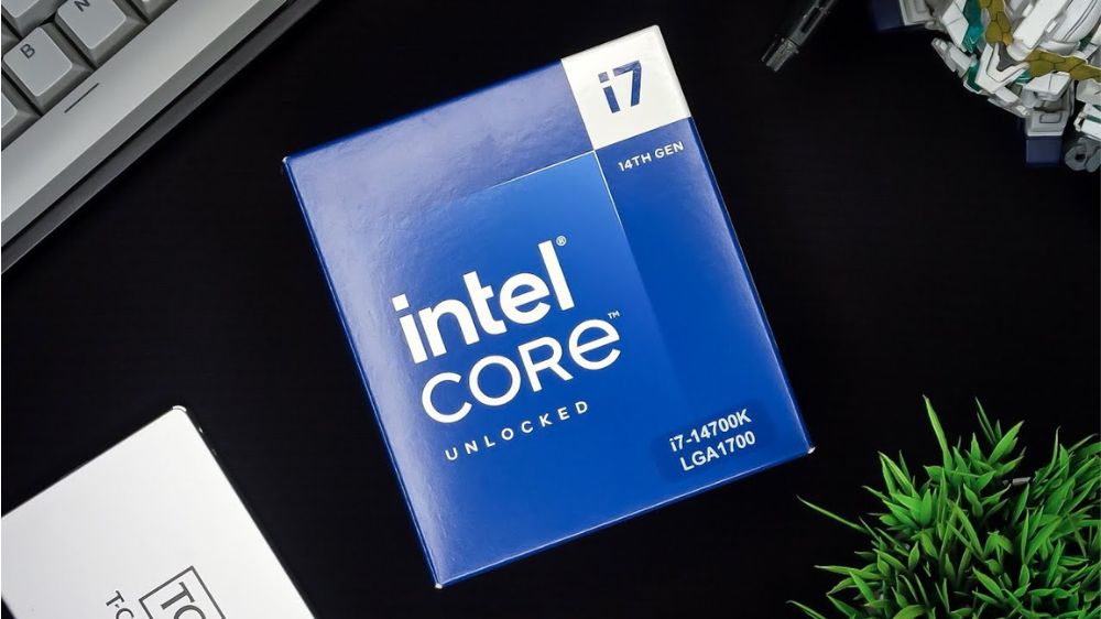جعبه آبی رنگ پردازنده اینتل مدل Core i7-14700K با لوگو Intel Core رویش، در حالی که روی میز مشکی رنگ قرار گرفته و یک کیبورد در سمت چپ پا و یک گیاه در سمت راست پایین آن قرار دارند