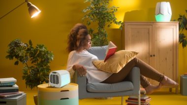 یک کولر رومیزی کوچک در اتاق و در حال زدن باد به سمت یک خانم روی مبل در حال کتاب خواندن
