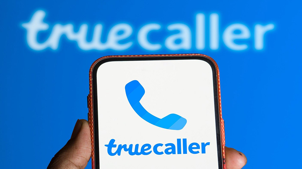 یک گوشی در حال نمایش آیکون برنامه Truecaller در دست یک نفر با لوگو برنامه در پسزمینه