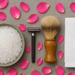 کاسه‌ای نمک بدن و تیغ اصلاح و برس اصلاح و حوله روی سطح که گلبرگ نیز اطراف آن پراکنده‌اند