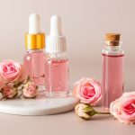 چند بطری گلاب و چند گل رز در یک پس‌زمینه صورتی