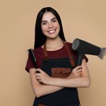 آرایشگری که لبخند می‌زند،‌دست به سینه ایستاده و یک شانه و سشوار در دست‌هایش دارد.