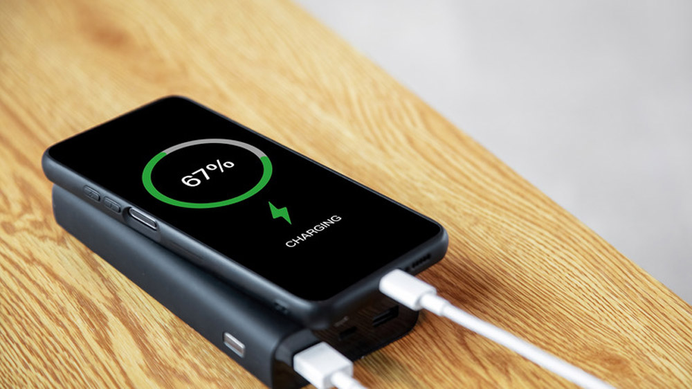 یک گوشی در حال شارژ متصل به پاوربانک با کابل سفید روی پس‌زمینه چوبی