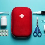 کیف کمک های اولیه قرمز با علامت صلیب سفید احاطه‌شده با اقلام کیت روی سطح آبی کمرنگ ساده