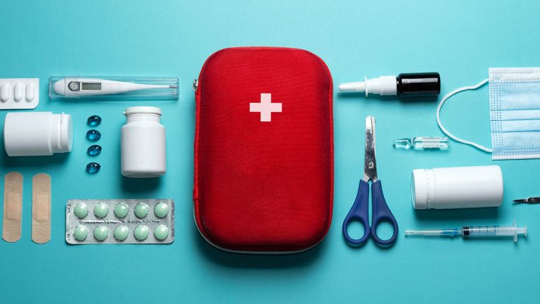 کیف کمک های اولیه قرمز با علامت صلیب سفید احاطه‌شده با اقلام کیت روی سطح آبی کمرنگ ساده