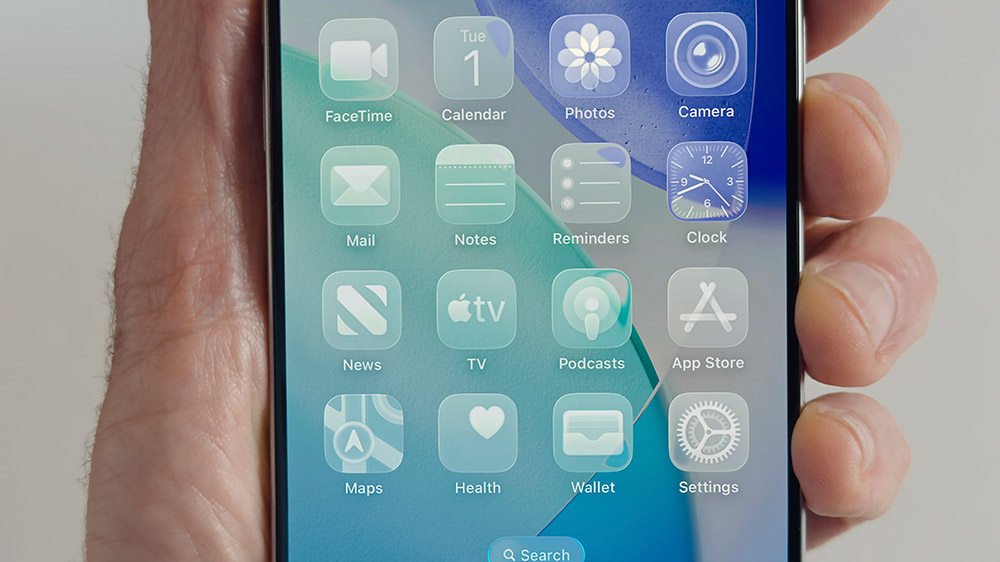 گوشی آیفون در حال نمایش منو اصلی iOS 26 با طراحی شیشهای در دست یک نفر