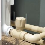 نمای کلوزاپ از یک رادیاتور سفید کنار دیوار با شیر ترموستات و لوله‌های پلاستیکی کرمی رنگ