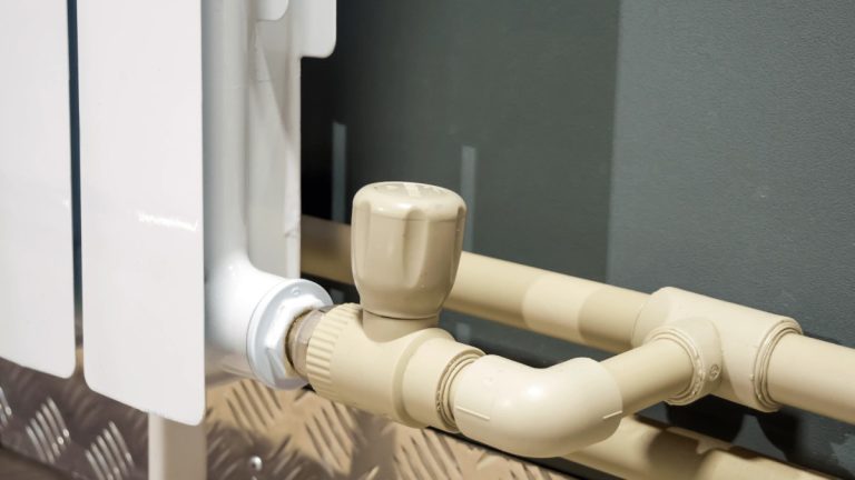 نمای کلوزاپ از یک رادیاتور سفید کنار دیوار با شیر ترموستات و لوله‌های پلاستیکی کرمی رنگ