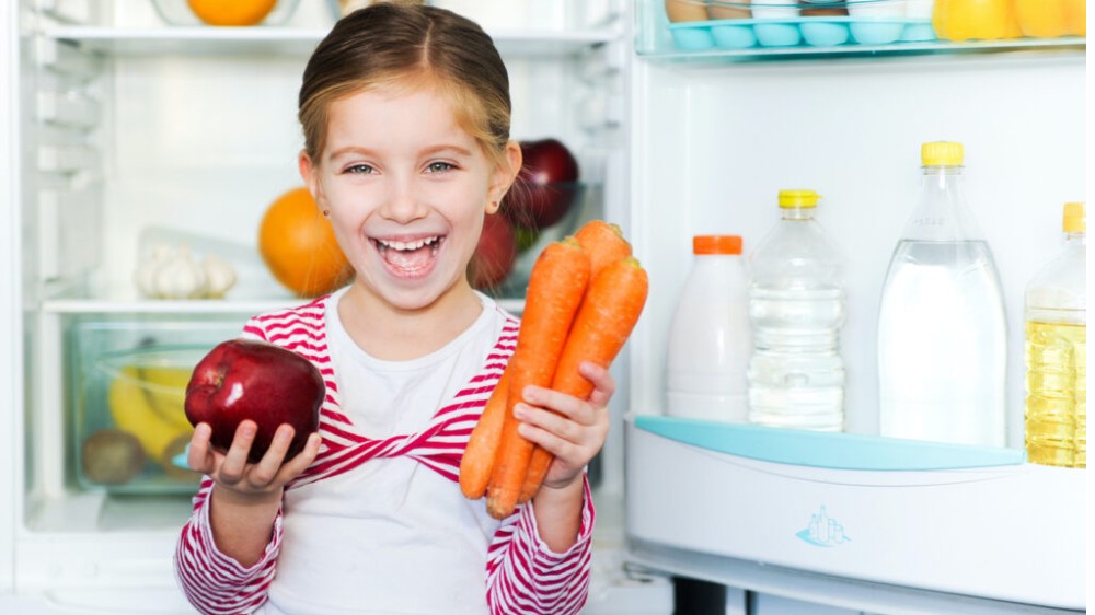 دختربچه‌ای شاد با سیب و هویج در دست، مقابل یخچال ایستاده است